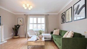 Una sala de estar con un sofá verde, un sillón blanco, una mesa de centro de madera y un ventanal con cortinas.