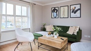 Una sala moderna con un sofá verde, un sillón blanco y una mesa de centro de madera. Hay tres cuadros enmarcados en la pared y una ventana con cortinas.