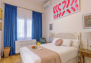 A hotel room with a large bed, a bedside table, a chair, a window with blue curtains, and artwork on the walls. - VILLA MARIA (Málaga)