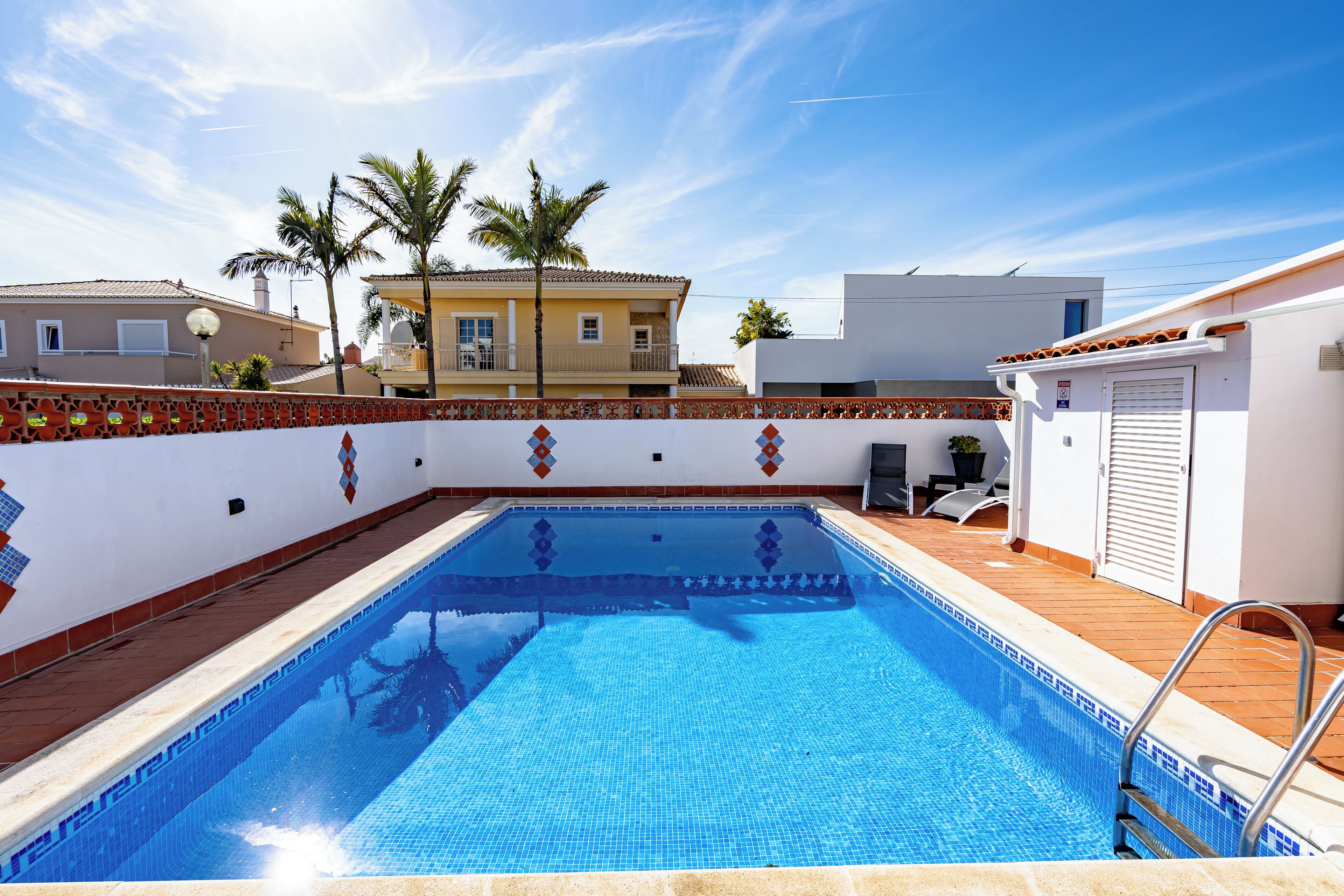 A swimming pool with a tiled edge, a tiled deck, and a white wall with a decorative border.
