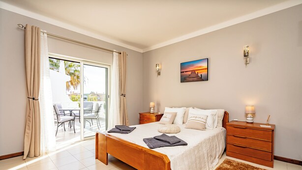 A bedroom with a bed, bedside tables, a nightstand, a lamp, a painting, and a view through a sliding glass door. - Algarve Charm with Private Pool, Garden & Spacious Area in Lagos (Lagos)