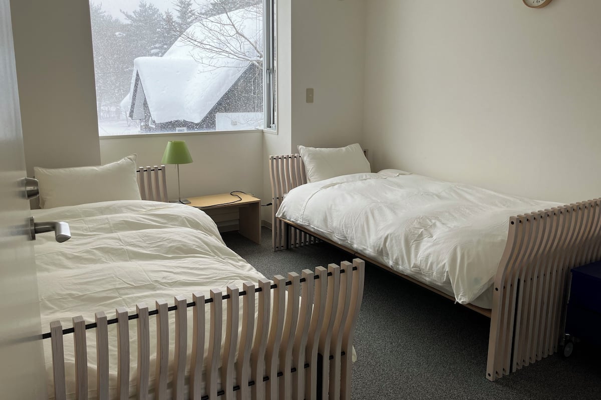 A room with two beds, a desk, a lamp, and a window with a view of trees and a snow-covered roof.