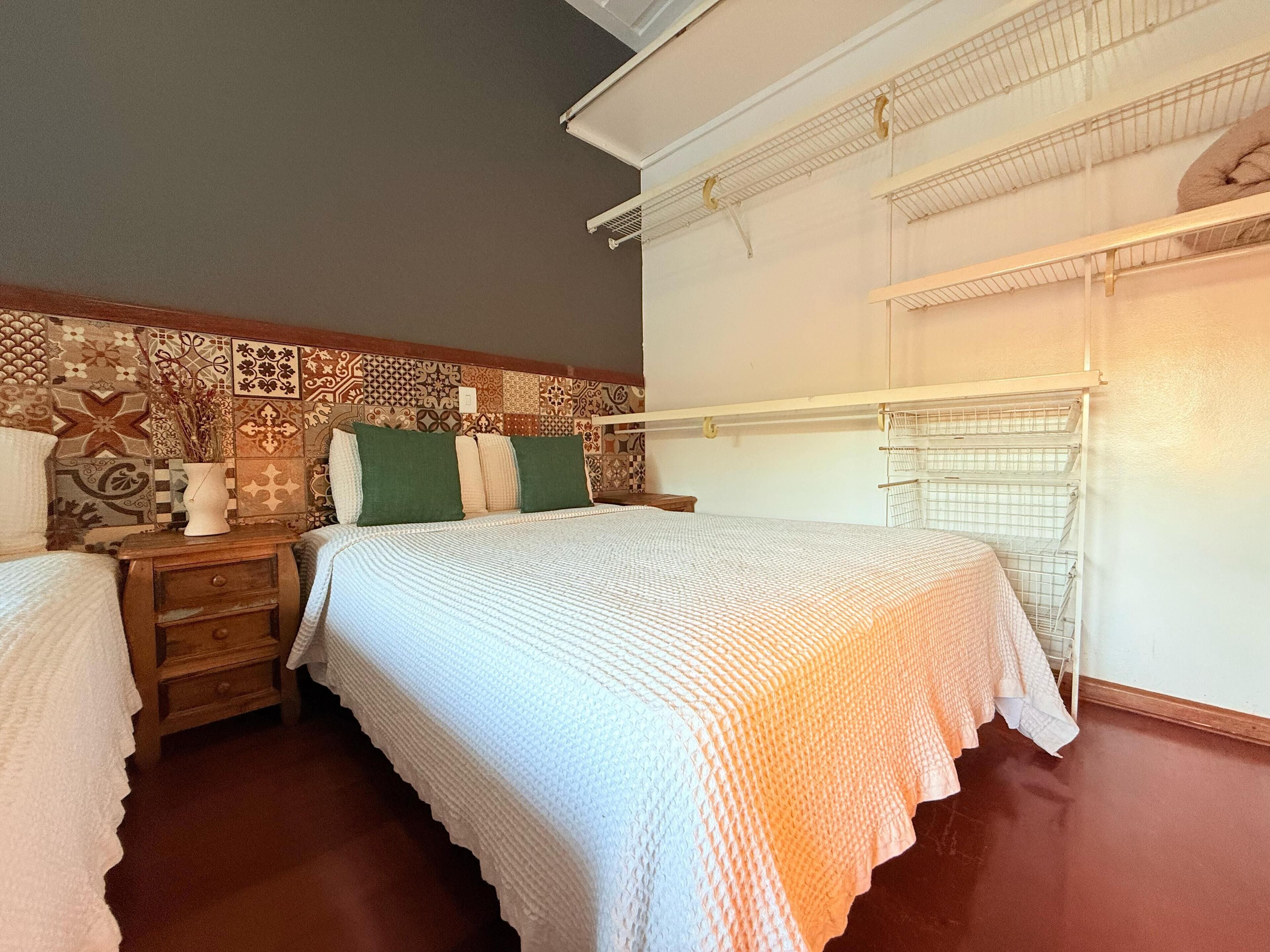 A bedroom with a patterned wall, a bed with white bedding, a nightstand, a dresser, and a metal shelf unit.