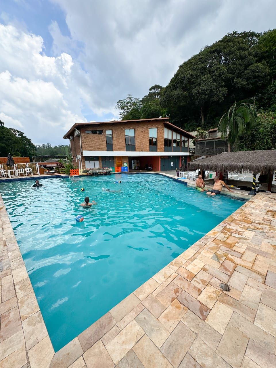 A swimming pool with people swimming and playing, surrounded by a building and trees.