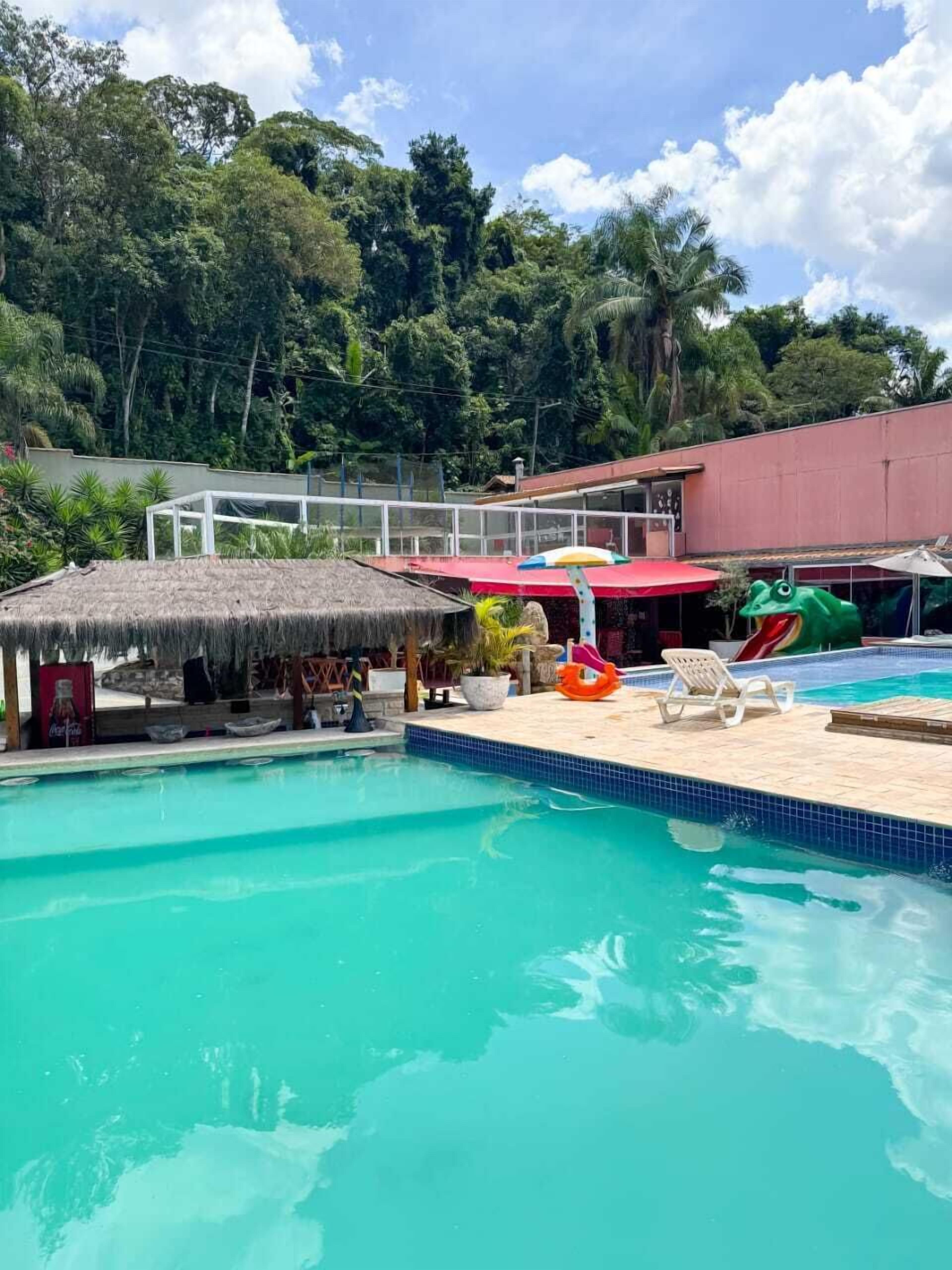 Piscina con sillas de descanso y un tobogán, rodeada de exuberante vegetación y un edificio con techo rojo.