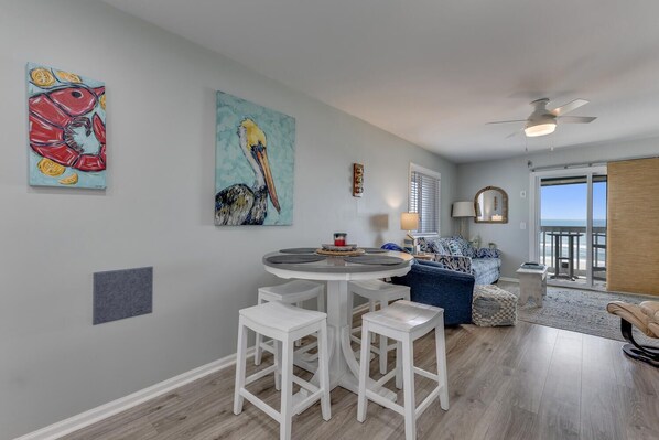 A modern living room with a round dining table, white bar stools, a blue sofa, and a view of the ocean through large sliding glass doors.