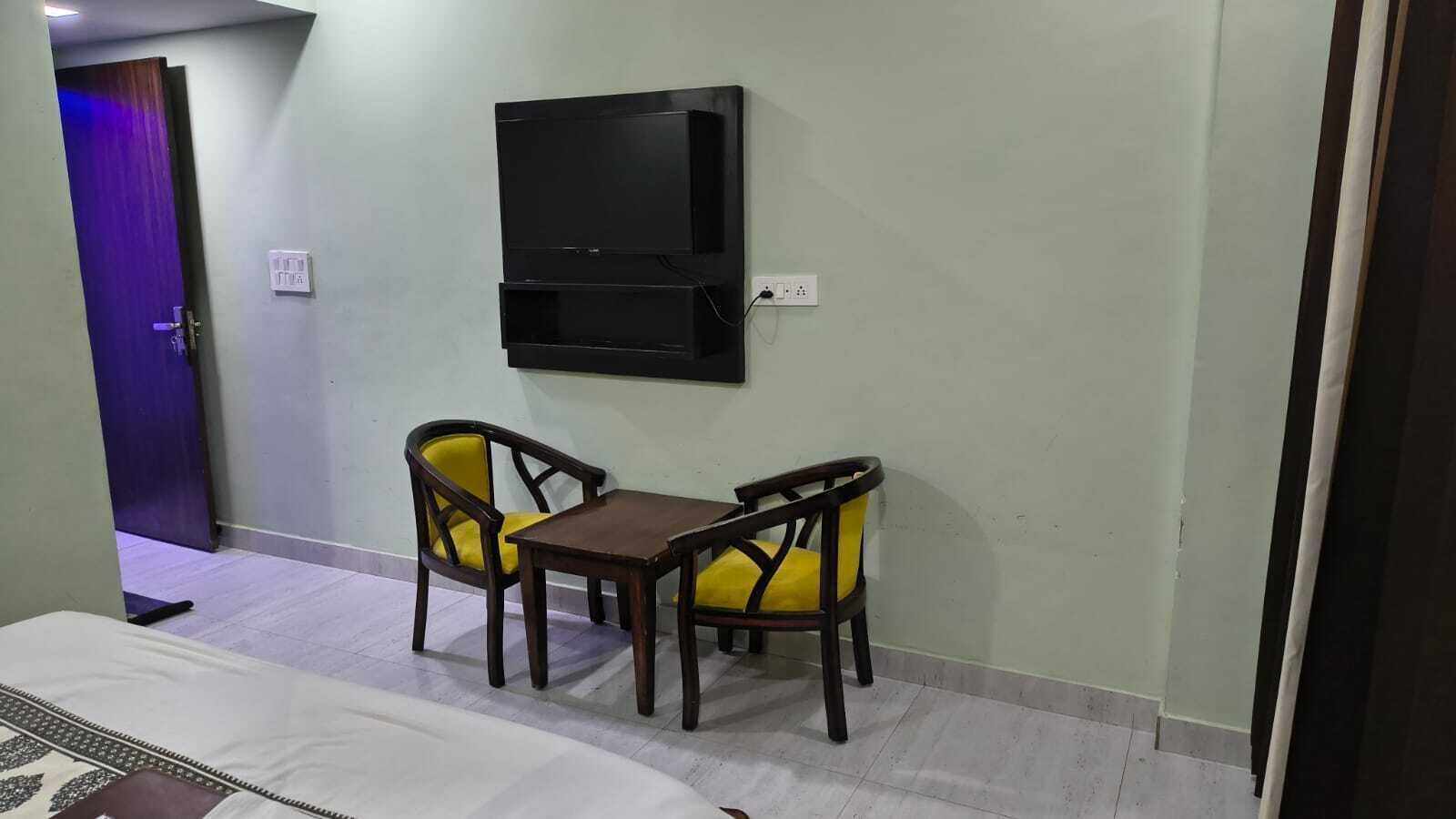 Una habitación de hotel con cama, una mesita, dos sillas amarillas y un televisor montado en la pared.