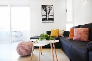 A modern living room with a black sofa, a round ottoman, a small white table with a plant, and a framed picture on the wall. - Intimate 1BR near Bograshov beach by HolyGuest (Tel Aviv-Yafo)