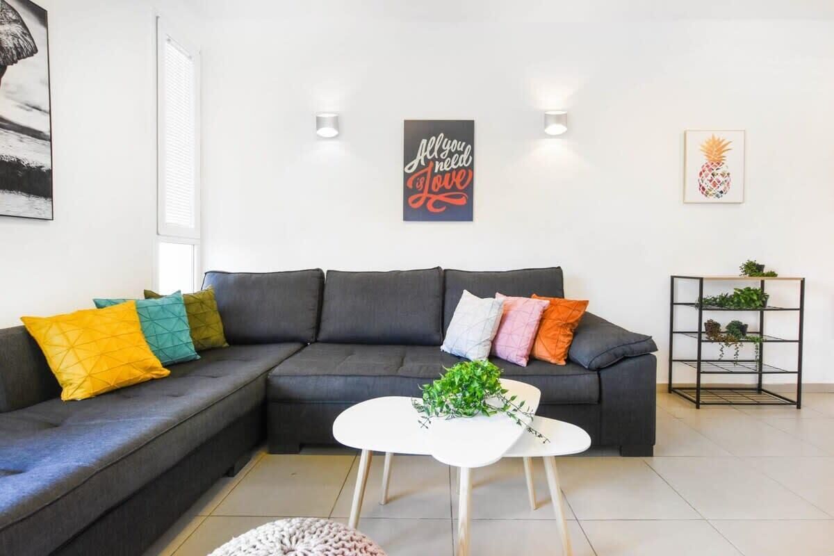 Un salon moderne avec un canapé d’angle, une table basse blanche et des coussins décoratifs. Des lampes murales et une œuvre d’art encadrée ornent le mur au-dessus du canapé.