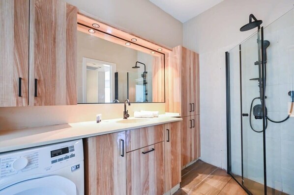 A modern bathroom with wooden cabinetry, a built-in washing machine, a glass shower enclosure, and a large mirror.