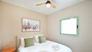 Una camera d'albergo con un letto, una ventola a soffitto, un quadro appeso al muro, una finestra e un tavolino.