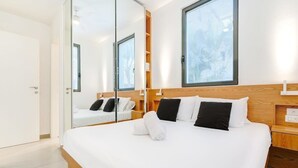A modern hotel room with a large bed, a wooden headboard, a bedside table, and a mirror reflecting the room.