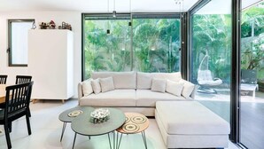 A modern living room with a large glass wall, a beige sofa, a dining table, and a small coffee table.