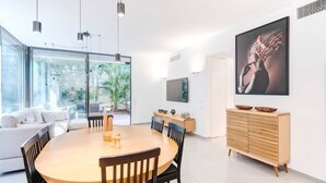 A modern dining area with a wooden table and chairs, a white sofa, a wooden cabinet with a flat-screen TV, and a large window with a view of greenery.
