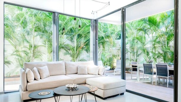 A modern living room with a beige sectional sofa, two round coffee tables, and a glass wall offering a view of lush greenery.