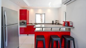 A modern kitchen with red cabinets, a stainless steel refrigerator, a coffee machine, and a toaster. There are three black stools at the counter. - Fragrant Flower 3BR by HolyGuest - Free Parking (Tel Aviv)