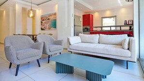 A modern living room with a sofa, two patterned chairs, a blue coffee table, and a red kitchen in the background. - Fragrant Flower 3BR by HolyGuest - Free Parking (Tel Aviv)
