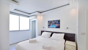 A modern hotel room with a large bed, white linens, a nightstand, and a painting of a pool on the wall. - Fragrant Flower 3BR by HolyGuest - Free Parking (Tel Aviv)