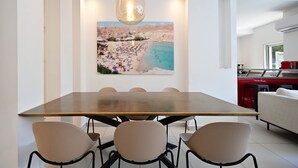 A dining area with a wooden table and beige chairs, a pendant light, and a beach scene painting on the wall. - Fragrant Flower 3BR by HolyGuest - Free Parking (Tel Aviv)