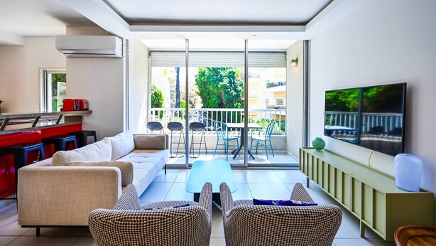 A modern living room with a sofa, coffee table, and wicker chairs. A TV is mounted on the wall, and a balcony with a view of the neighborhood is visible. - Fragrant Flower 3BR by HolyGuest - Free Parking (Tel Aviv)