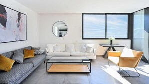 A modern living room with a sofa, coffee table, and a chair. There is a large window providing natural light and a view outside.