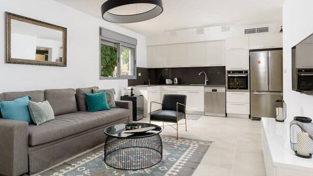 Een moderne woonkamer met een grijze bank, een zwarte salontafel en een kamerbreed tapijt. Een zwarte stoel staat nabij een witte keuken met roestvrijstalen apparatuur en witte kasten.