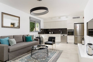 A modern living room with a grey sofa, black coffee table, and a patterned rug. A black chair is positioned near a white kitchen with stainless steel appliances and white cabinets.