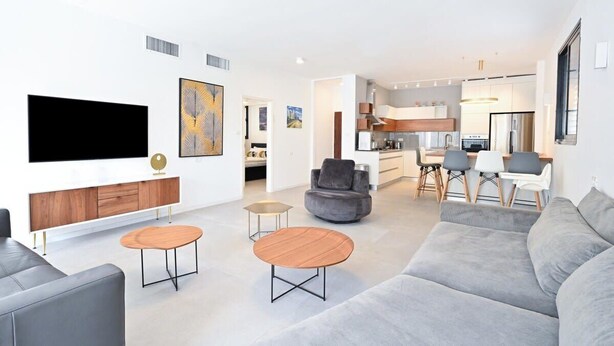 Un salon moderne avec un téléviseur à écran plat, un canapé gris, deux petites tables rondes et un fauteuil gris.