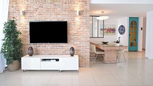Un salon avec un mur en briques, un téléviseur à écran plat, un meuble de rangement blanc, un coin repas avec une porte de couleur turquoise et une plante en pot.
