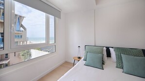 Een slaapkamer met een groot raam, een bed met groene kussens en een klein nachtkastje met een lamp.