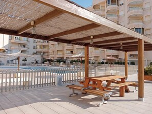 A covered outdoor area with picnic tables, a pool, and a white picket fence.