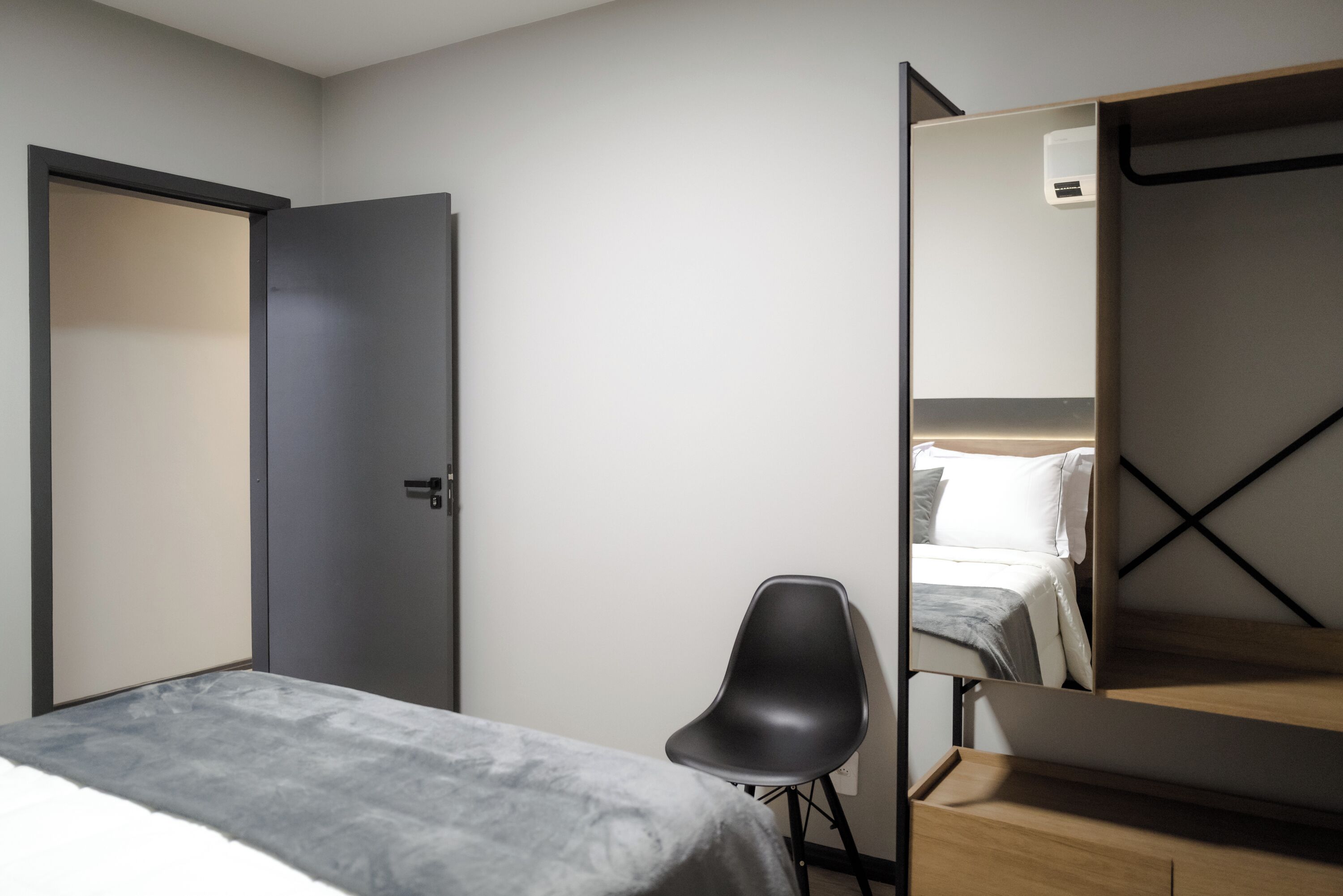 Ein modernes Hotelzimmer mit Bett, Sessel, Kleiderschrank und Spiegel, der das Bett und einen Nachttisch reflektiert.
