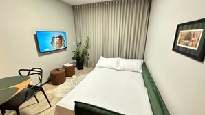Una camera d'albergo con un letto, una televisione a muro, una scrivania con una sedia, una pianta in vaso e un quadro incorniciato.