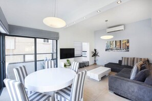 A modern living room with a round dining table, striped chairs, a grey sofa, a flat-screen TV, and a large window with blinds.