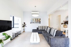 A modern living room with a large flat-screen TV, a grey sectional sofa, a white coffee table, and a large window. - Zebra's Stripe by HolyGuest (Tel Aviv)