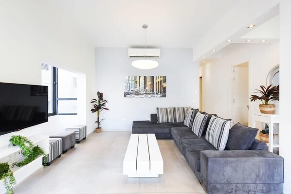 A modern living room with a large flat-screen TV, a grey sectional sofa, a white coffee table, and a large window. - Zebra's Stripe by HolyGuest (Tel Aviv)