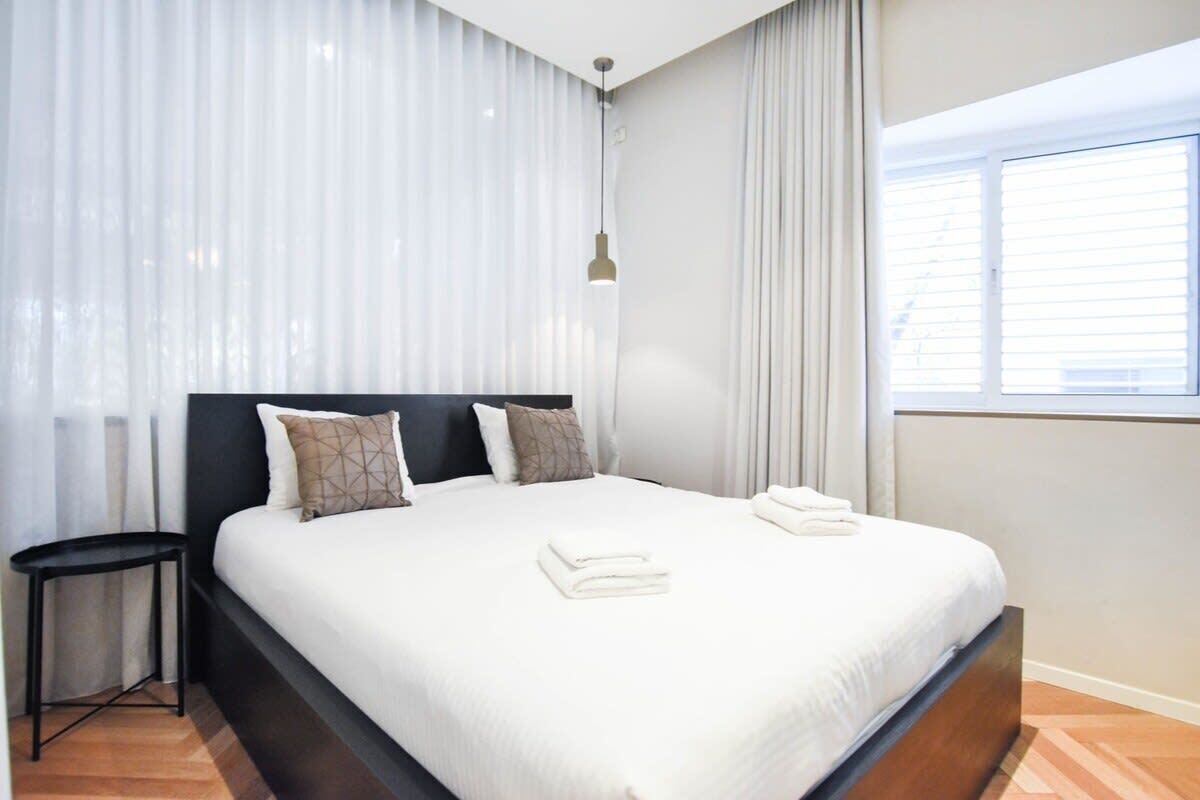A modern hotel room with a large bed, a bedside table, and a window with blinds.