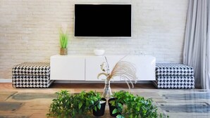 A modern living room with a white brick wall, a mounted TV, two patterned ottomans, a white cabinet, and a central plant arrangement.