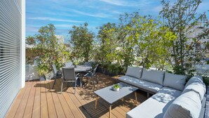 Une terrasse extérieure avec une plateforme en bois, un canapé gris, une table à manger avec des chaises et des plantes en pot.