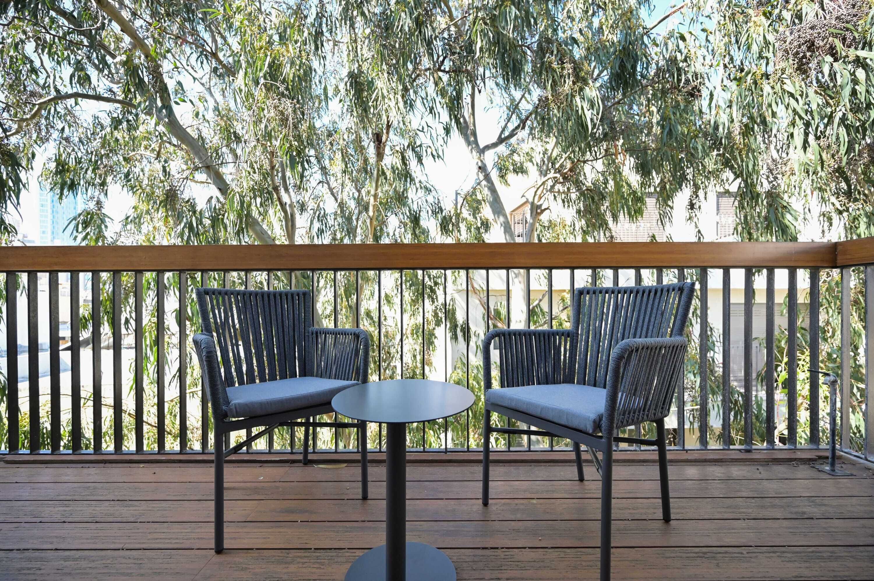 A balcony with two chairs and a small round table, surrounded by a wooden railing and lush green trees.