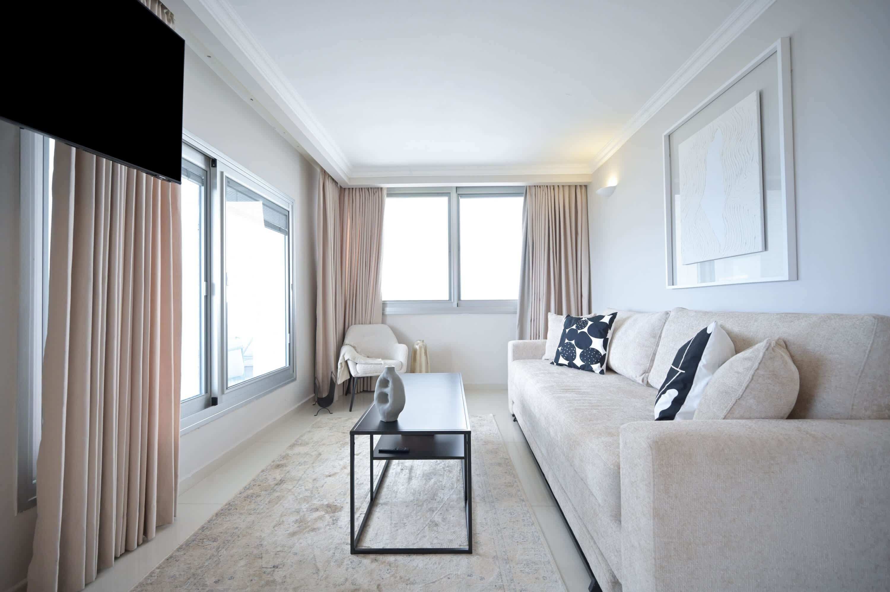 A modern living room with a sofa, a coffee table, and a chair. There are large windows with curtains, a flat-screen TV, and a framed artwork on the wall.