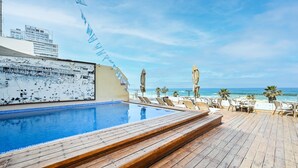 Un área de piscina con terraza de madera, sillas de descanso y vista al océano.