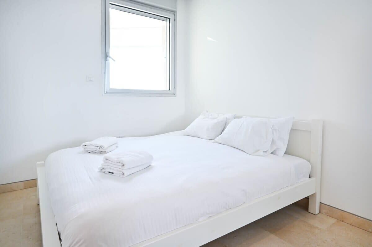 A white single bed with a headboard, a window above it, and a neatly folded white towel on the bed.