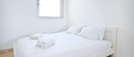 A white single bed with a headboard, a window above it, and a neatly folded white towel on the bed.