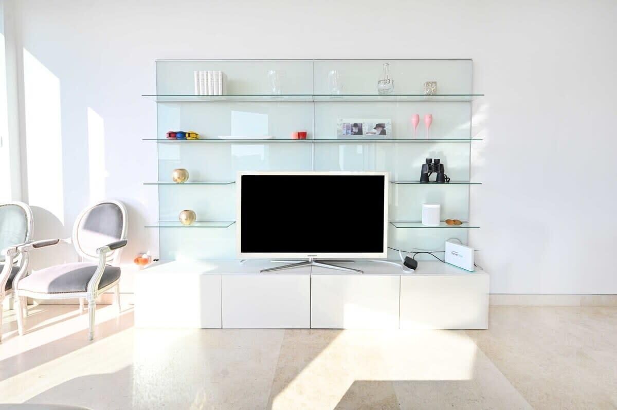 Een moderne woonkamer met een glazen tv-meubel, witte meubels en een boekenkast met diverse spullen.