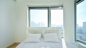 A modern hotel room with a large bed, white bedding, and a city view through large windows.