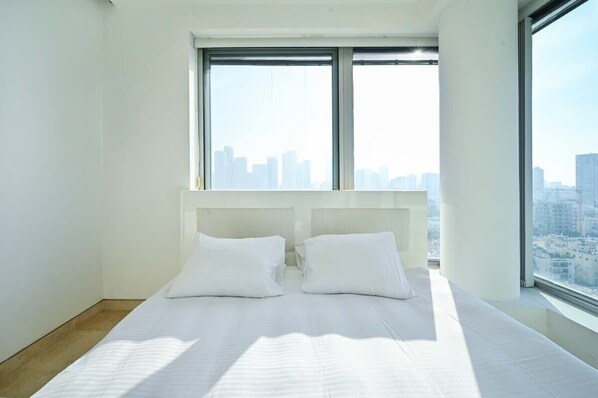 Habitación de hotel moderna con una cama grande, ropa de cama blanca y vistas a la ciudad a través de amplios ventanales.