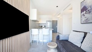 A modern living room with a dark grey sofa, a white coffee table, and a large flat-screen TV mounted on a wooden wall. - Cozy 1BR Apartment in Mendele by HolyGuest (Tel Aviv)