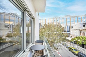 Un balcón con una mesa, sillas, un ventanal y vista a una calle con edificios y automóviles estacionados.