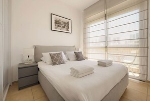 A modern bedroom with a large bed, a nightstand, a bedside table, a framed picture on the wall, and a view through the window.